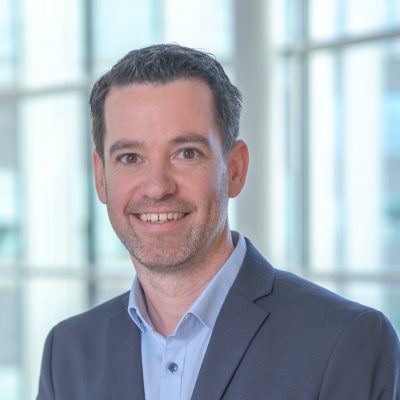 Man in een blauw colbert en lichtblauw overhemd glimlacht naar de camera in een moderne, lichte kantooromgeving met grote ramen op de achtergrond