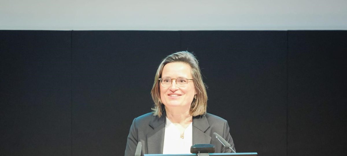 Vrouw in een donker colbert geeft een presentatie achter een spreekgestoelte, met een glimlach, in een conferentiezaal.