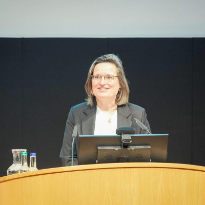 Vrouw in een donker colbert geeft een presentatie achter een spreekgestoelte, met een glimlach, in een conferentiezaal.