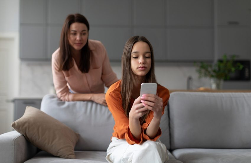 Meisje zit op de bank met haar smartphone in haar handen. Haar moeder kijkt over haar schouder mee.